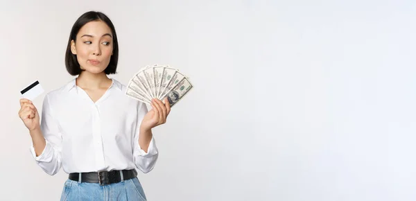 Εικόνα ασιάτισσας που κοιτάζει τα χρήματα των δολαρίων, κρατώντας την πιστωτική κάρτα σε ένα άλλο χέρι, σκέφτεται, στέκεται πάνω από το λευκό φόντο — Φωτογραφία Αρχείου