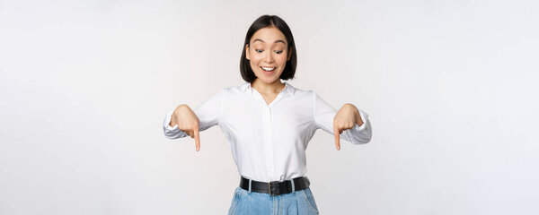 Portrait of happy asian woman pointing fingers down and looking below at advertisement, showing info banner, advertising, standing over white background