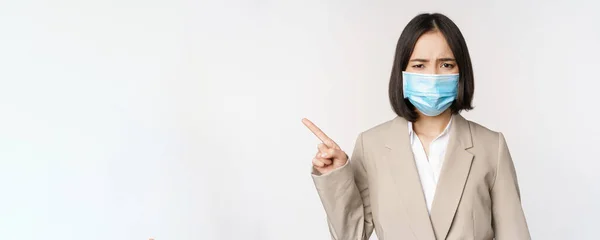 Coronavirus och arbetskoncept. Frustrerad asiatisk affärskvinna i medicinsk ansiktsmask, pekar finger vänster och klagar, står över vit bakgrund — Stockfoto