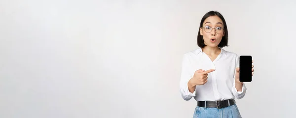 Entusiasta joven asiática mujer apuntando con el dedo a la pantalla del teléfono inteligente, mostrando publicidad en el teléfono móvil, fondo blanco — Foto de Stock