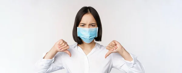 Portret Azjatki w medycznej masce twarzy z kciukami w dół, rozczarowany, zmęczony wyraz twarzy, stojący na białym tle — Zdjęcie stockowe