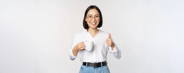 Gülümseyen Asyalı kız elinde kupa, beyaz kupa ve baş parmaklar yukarı, içki, kahve ya da çay öneriyor, beyaz arka planda duruyor. — Stok fotoğraf