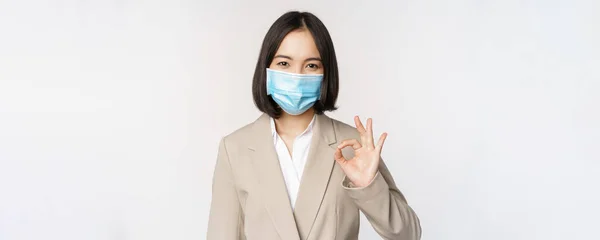 Coronavirus and workplace concept. Image of asian saleswoman, company worker in medical mask showing okay sign, smiling pleased, white background — Stock Photo, Image