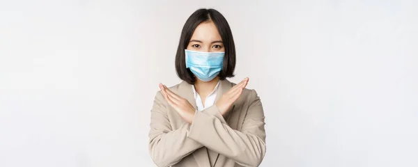 Concepto de Coronavirus y lugar de trabajo. Imagen de mujer de negocios asiática, mujer en máscara facial médica muestra parada, gesto de prohibición cruzada, se para sobre fondo blanco —  Fotos de Stock