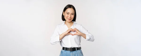 Liefde gebaar. Mooie aziatische jonge vrouw, met hart gebaar en glimlach, uitdrukkelijke zorg en genegenheid, staande over witte achtergrond — Stockfoto