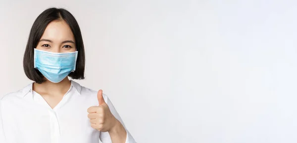 Ковид и концепция здравоохранения. Портрет азиатки, носящей медицинскую маску и показывающей большие пальцы, стоящей на белом фоне — стоковое фото