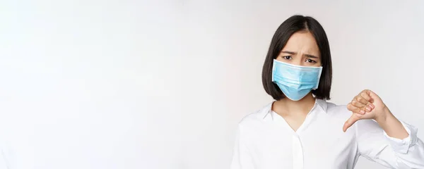 Porträt einer asiatischen Frau mit medizinischer Gesichtsmaske, die Daumen nach unten zeigt, mit enttäuschtem, müdem Gesichtsausdruck, der über weißem Hintergrund steht — Stockfoto