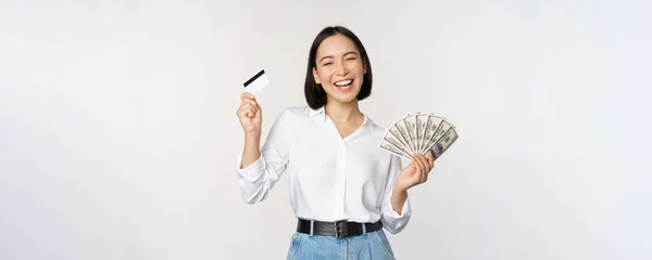 Mulher coreana feliz segurando cartão de crédito e dinheiro dólares, sorrindo e rindo, posando contra fundo estúdio branco — Fotografia de Stock