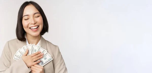 Glad asiatisk affärskvinna innehar kontanter, kramar dollar pengar och ler, står över vit bakgrund i kostym — Stockfoto