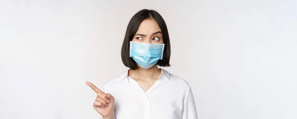Bild av ung asiatisk kvinna i medicinsk ansiktsmask, pekar vänster och ser med misstänksam förvirrad uttryck, står över vit bakgrund — Stockfoto