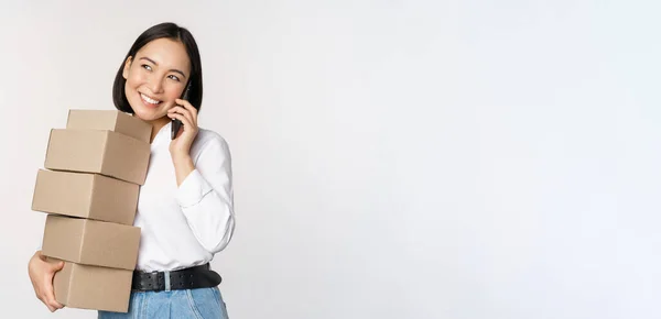 Imagem de jovem mulher de negócios asiática atender telefonema enquanto carrega caixas para entrega, posando contra fundo branco — Fotografia de Stock