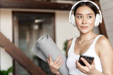 Genç Asyalı kadın kulaklıkla evde duruyor, müzik dinliyor ve paspasla egzersiz yapıyor, yoga yapıyor ya da oturma odasında meditasyon yapıyor.