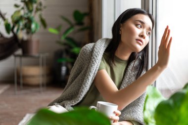 Üzgün Koreli kız pencereye dokunuyor ve hasretli üzgün yüzüyle dışarı bakıyor, düşünceli yüzüyle uzaktan bakıyor.