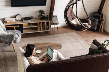 Evde dizüstü bilgisayarla çalışan Asyalı bir kadının portresi. Kanepede oturup dinleniyor. Oturma odasında bilgisayar olan Koreli kız.