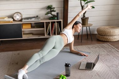 Genç Asyalı kadın evde paspasın üzerinde antrenman yapıyor, spor eğitmenini dizüstü bilgisayarda takip ediyor, kolunu uzatıyor ve kalas yapıyor.