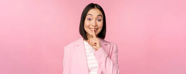 Silencio, concepto tabú. Retrato de una mujer de negocios asiática mostrando un gesto de silencio, un signo de shhh, presiona el dedo contra los labios, de pie sobre un fondo rosa en traje — Foto de Stock