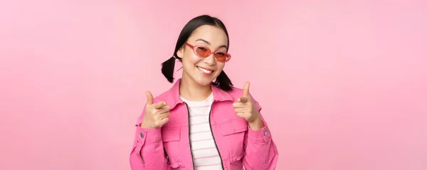 Close up portret nowoczesnej azjatyckiej dziewczyny w okularach przeciwsłonecznych uśmiecha, wskazując palcami na aparat, chwalić, zapraszając lub komplement, stojąc nad różowym tle — Zdjęcie stockowe