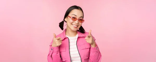 Primer plano retrato de la chica asiática moderna en gafas de sol sonriendo, señalando los dedos a la cámara, alabanza, invitando o elogiando, de pie sobre fondo rosa — Foto de Stock