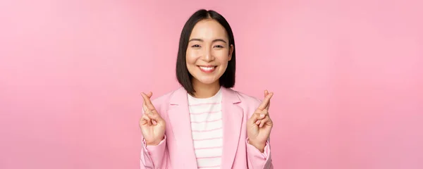 Feliz asiático empresária cruz dedos para boa sorte, desejando, orando e esperando, sorrindo para a câmera, de pé em terno sobre rosa fundo — Fotografia de Stock