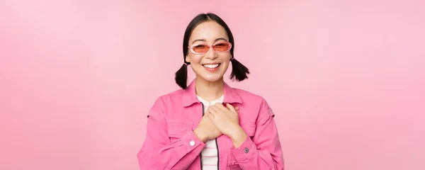 Usmívající se mladá Asiatka ve slunečních brýlích vypadá vděčně, drží se za ruce na srdci, polichocen, ocení smth, stojící nad růžovým pozadím — Stock fotografie