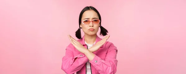 Ritratto di ragazza coreana in occhiali da sole alla moda, imbronciato deluso, mostrando stop, gesto di rifiuto, segno croce, in piedi su sfondo rosa — Foto Stock
