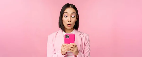 Portrait de femme d'affaires asiatique au visage surpris, utilisant une application smartphone, portant un costume d'affaires. Fille coréenne avec téléphone mobile et expression du visage excité, fond rose — Photo