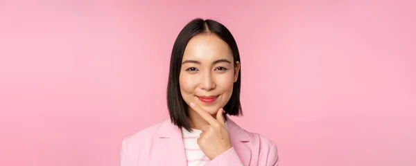 Крупним планом портрет усміхненої азіатської робочої леді в костюмі, бізнес-леді виглядає вдумливою, думає або вирішує smth, стоїть над рожевим тлом — стокове фото