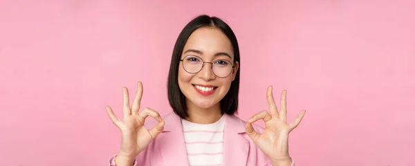 Detail portrét ohromen korporátní žena, asijské obchodní dáma v brýlích, ukazující v pořádku znamení, při pohledu na kameru, doporučující, růžové pozadí — Stock fotografie
