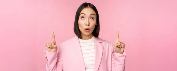 Ενθουσιαστικός εταιρικός εργαζόμενος, ασιάτισσα επιχειρηματίας δείχνει τα δάχτυλα και χαμογελά, δείχνει διαφήμιση, λογότυπο, στέκεται πάνω από ροζ φόντο — Φωτογραφία Αρχείου