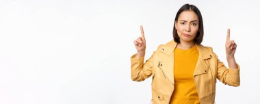 Şüpheci Asyalı kadın parmak kaldırıyor, kaşlarını çatıyor, onaylamıyor, şikâyet ediyor, beyaz arka planda şüpheli duruyor.