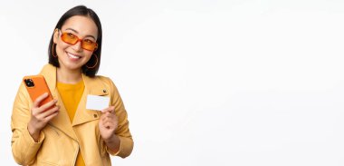 Online alışveriş ve teslimat konsepti. Şık elbiseli mutlu Koreli kız, elinde kredi kartı ve akıllı telefon, gülüyor ve gülümsüyor, beyaz arka planda duruyor.