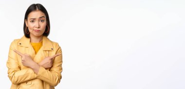 Kararsız Asyalı kız tasviri, parmakları yana, solu ve sağı işaret ediyor, varyantı seçiyor, karar veriyor, beyaz arka planın üzerinde duruyor.