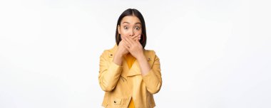 Şok olmuş Asyalı bir kız görüntüsü ağzını kapatır, dudaklarını kapatır ve nutku tutulur, korkmuş yüz ifadesi, beyaz arka planda durur.