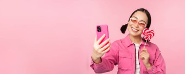 Şekerle, lolipop tatlılarıyla selfie çeken, gülümseyen, mobil uygulamayla fotoğraf çeken pembe arka planda duran şık, mutlu Asyalı kızın portresi.