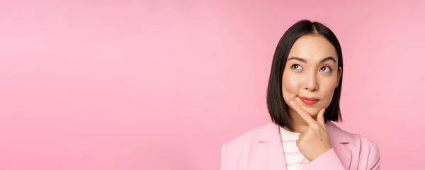 Primer plano retrato de joven mujer de negocios asiática pensando, sonriendo pensativo y mirando a la esquina superior izquierda, de pie sobre fondo rosa —  Fotos de Stock