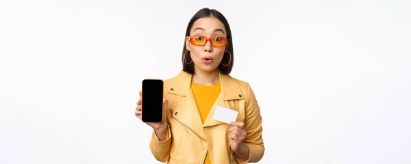 Compras en línea y concepto de personas. Mujer asiática con estilo mostrando la pantalla del teléfono móvil y la tarjeta de crédito, aplicación de teléfono inteligente, de pie sobre fondo blanco — Foto de Stock