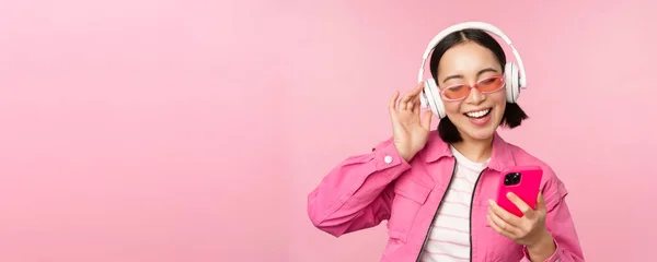 Elegante chica asiática bailando con smartphone, escuchando música en auriculares en la aplicación del teléfono móvil, sonriendo y riendo, posando sobre fondo rosa — Foto de Stock