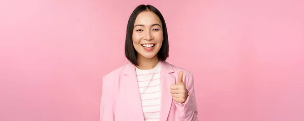 Porträt einer asiatischen Geschäftsfrau, die zufrieden lächelt, Daumen nach oben zeigt, Lob lobt, gefällt und billigt, im Anzug vor rosa Hintergrund steht — Stockfoto