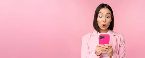 Портрет азиатской бизнесвумен с удивленным лицом, с помощью приложения для смартфонов, в деловом костюме. Корейская девушка с мобильным телефоном и возбужденное выражение лица, розовый фон — стоковое фото