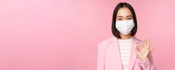 Porträtt av asiatisk affärskvinna i medicinsk ansiktsmask, visar okej tecken, bär kostym, arbetsregler under covid-19 pandemi, står över rosa bakgrund — Stockfoto