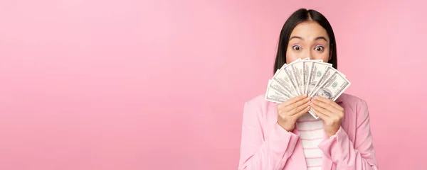 Szczęśliwy Azji lady w garniturze gospodarstwa pieniądze, dolary z zadowolony wyraz twarzy, stojąc na różowym tle — Zdjęcie stockowe
