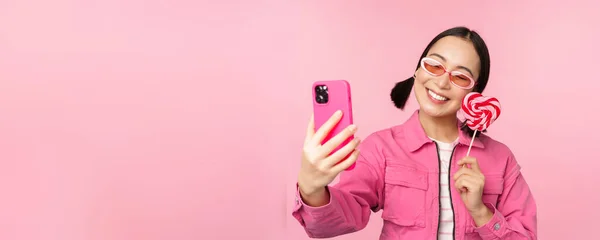 Portret van stijlvol, gelukkig aziatisch meisje dat selfie maakt met snoep, lolipop snoep en glimlach, foto 's maakt met mobiele app, over een roze achtergrond staat — Stockfoto