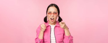 Image of angry, pissed off korean adult female model, shaking fists and shouting, screaming outraged, standing over pink background