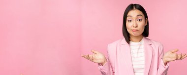 Kafası karışmış Asyalı iş kadınının portresi omuzlarını silkiyor, budala ve şaşkın görünüyor, bilemiyorum, bilemiyorum, pembe arka planda duran ofis elbisesinin içinde.