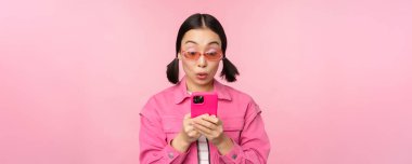 Akıllı telefon kullanan Asyalı bir kızın portresi. Kadın cep telefonuna bakıyor, uygulamayı tarıyor, pembe arka planda duruyor.