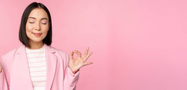 Sakin olun. Gülümseyen genç Asyalı kadın meditasyon yapıyor, yoga yapıyor, iş yerinde farkındalık ve gevşeme gösteriyor, nefes alıyor, keyifli bir şekilde havayı soluyor, pembe arka planda duruyor.