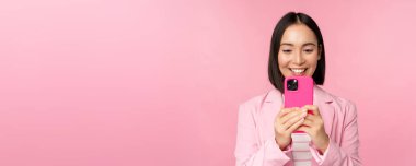 Gülümseyen Asyalı takım elbiseli bir kadının resmi, akıllı telefon uygulamasını izliyor, cep telefonu uygulamasını kullanıyor, pembe arka planda duruyor.