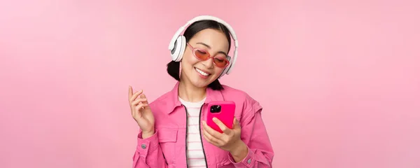 Стильна азіатська дівчина танцює зі смартфоном, слухаючи музику в навушниках на мобільному телефоні, посміхаючись і сміючись, позує на рожевому фоні — стокове фото