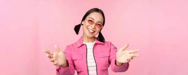 Chica coreana con estilo feliz en gafas de sol, llegar a las manos, recibir, sonreír amable y darle la bienvenida, de pie sobre el fondo rosa —  Fotos de Stock