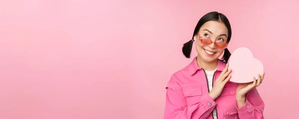 Imagen de elegante novia asiática en gafas de sol, adivinando lo que hay dentro de la caja de regalo, regalo en forma de corazón, de pie sobre fondo rosa — Foto de Stock